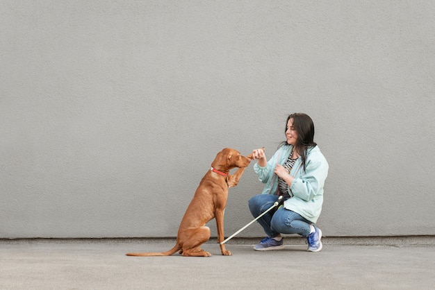 A woman is holding a leash attached to a medium-sized dog. She is standing calmly while the dog is sitting patiently on the floor. A guest is approaching them with a neutral expression.